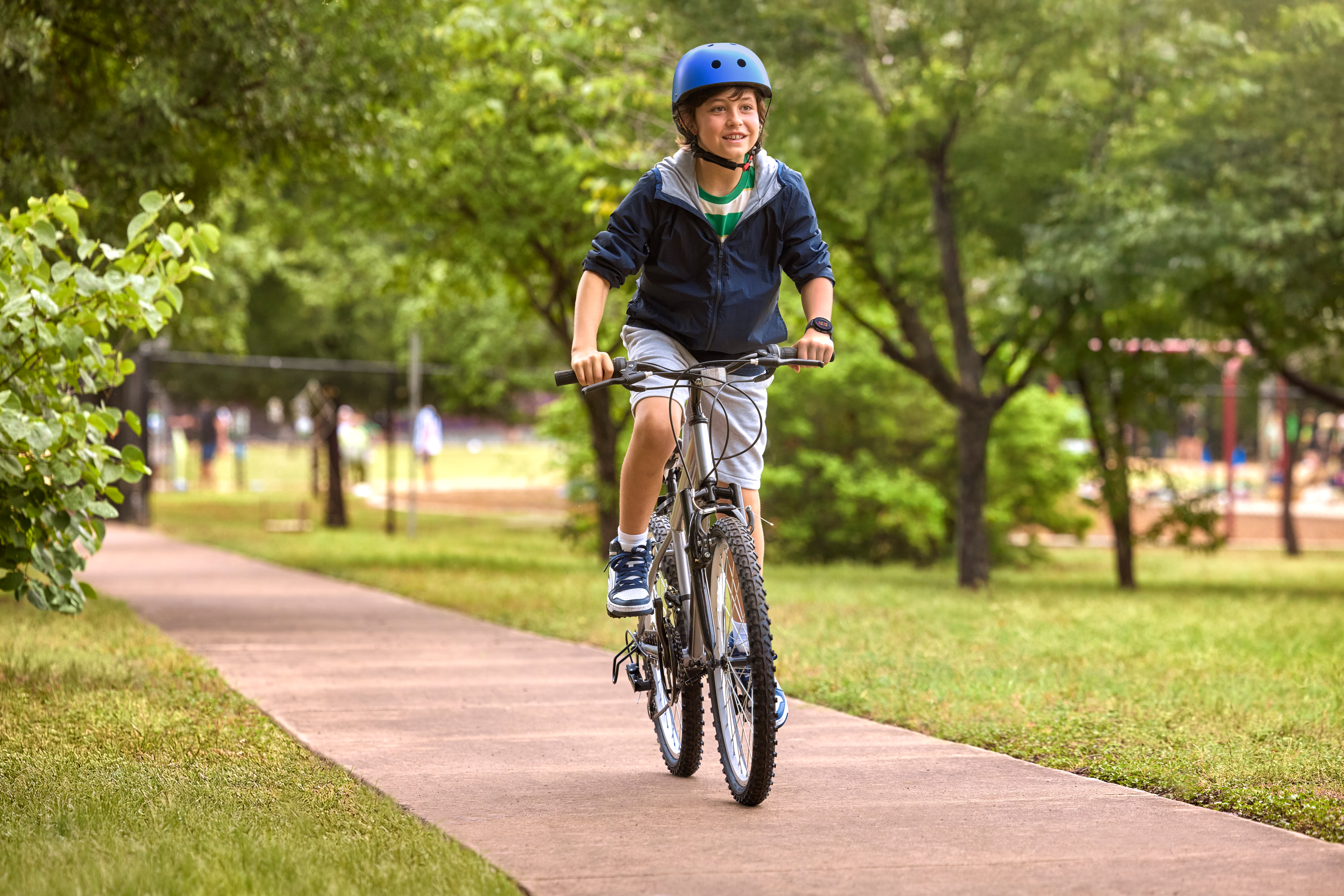 Ein Junge fährt Fahrrad auf einem Weg. Er trägt einen blauen Helm und eine Jacke. Grüne Bäume und Gras bilden den Hintergrund.