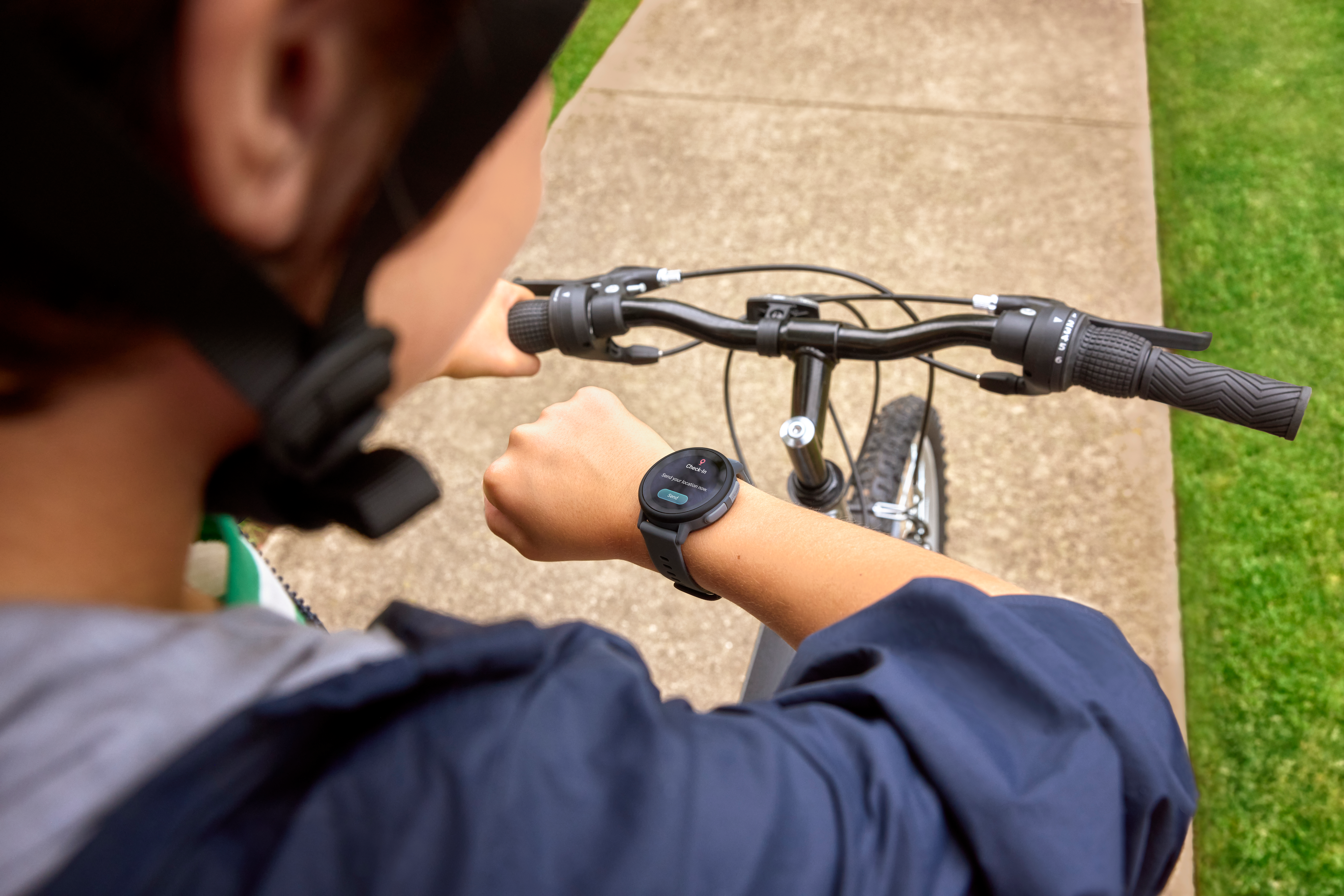 Eine Person auf einem Fahrrad schaut auf eine Smartwatch. Die Szene zeigt ein Fahrrad, einen Betonweg und grünes Gras.