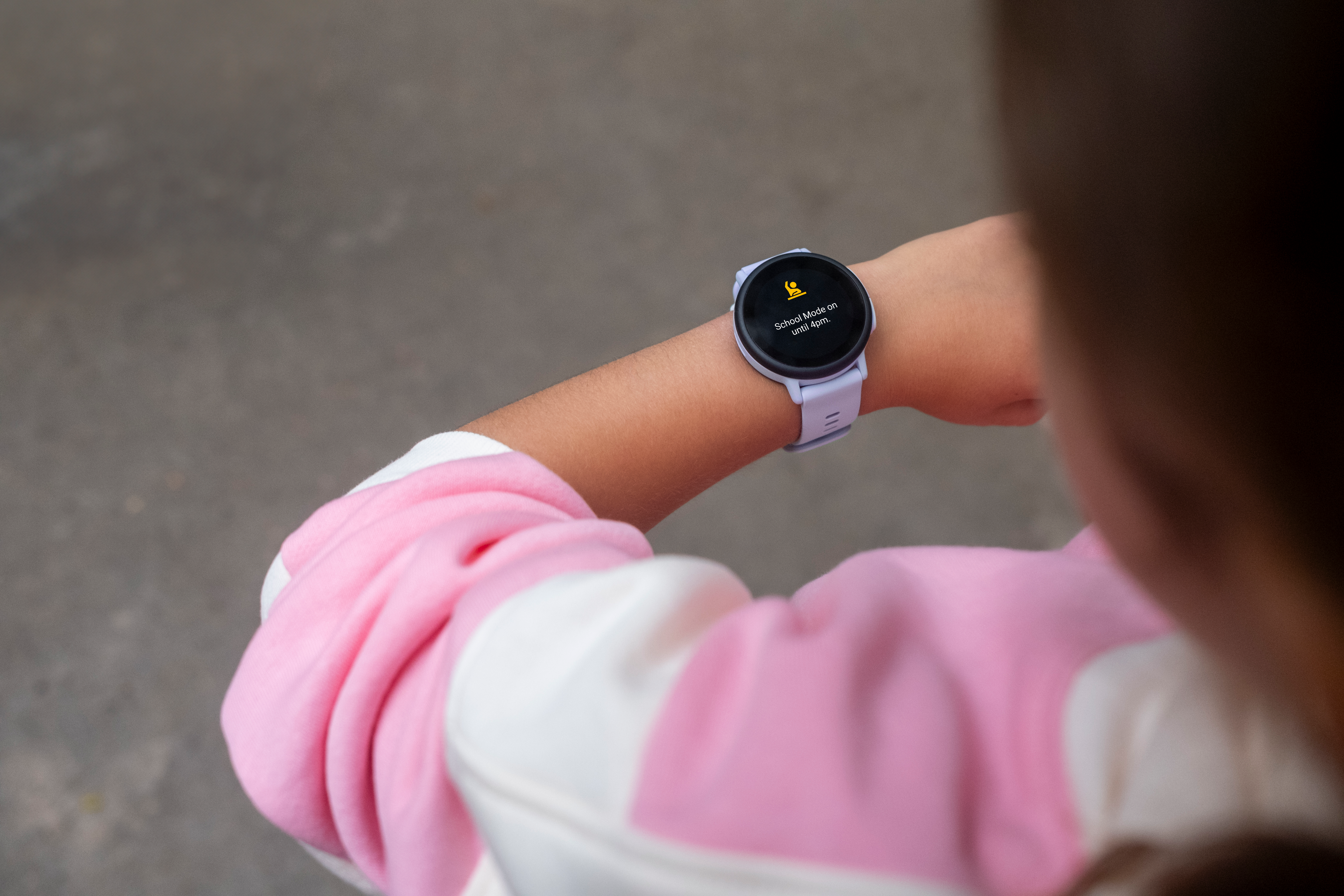 Der Arm eines Kindes mit einer Smartwatch, die 'Schulmodus aktiviert' anzeigt. Das Kind trägt ein rosa-weißes Oberteil. Grauer Hintergrund.