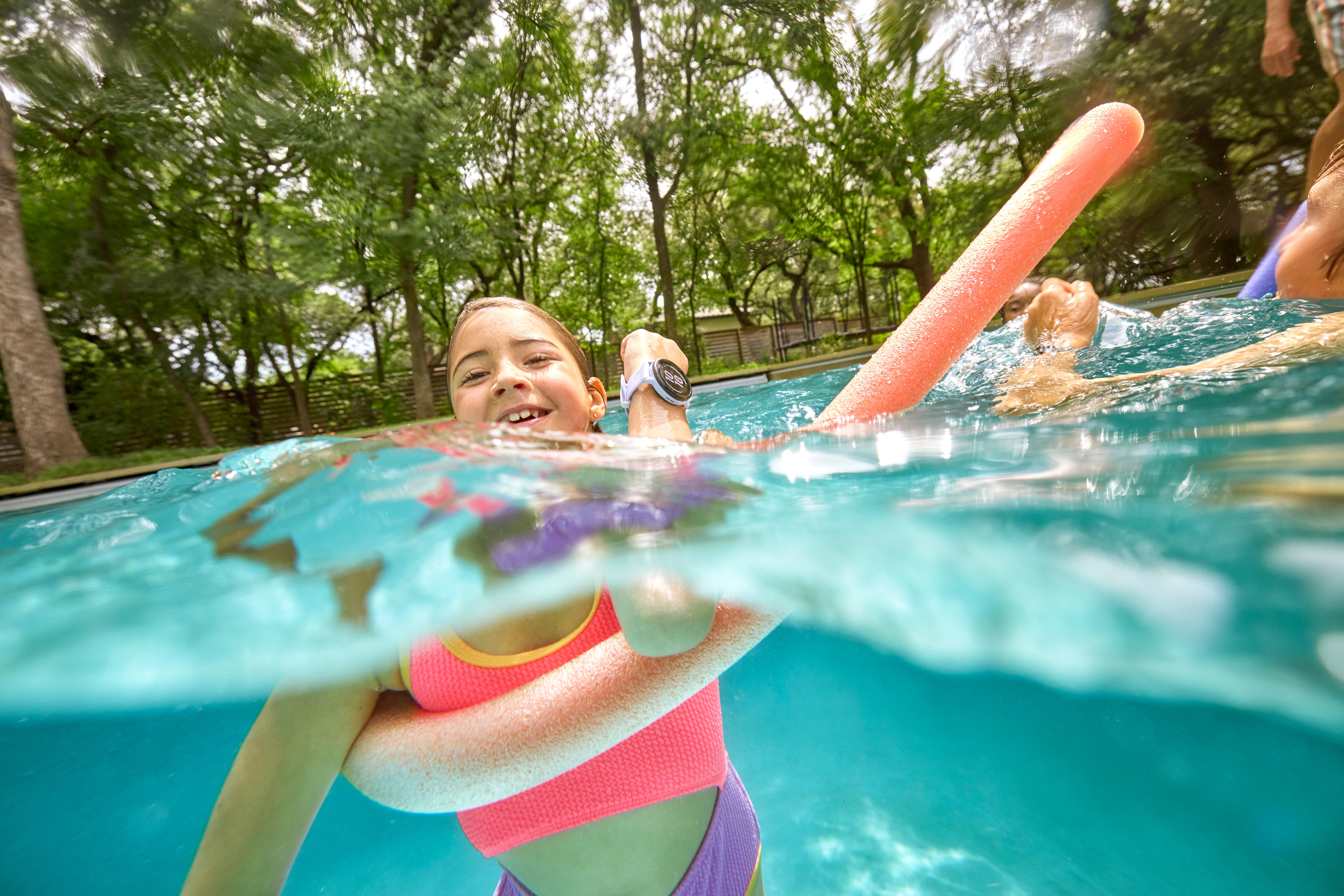 Ein Mädchen in einem Pool mit pinkfarbenem Oberteil und lila Unterteil, das eine pinkfarbene Poolnudel hält. Bäume im Hintergrund.