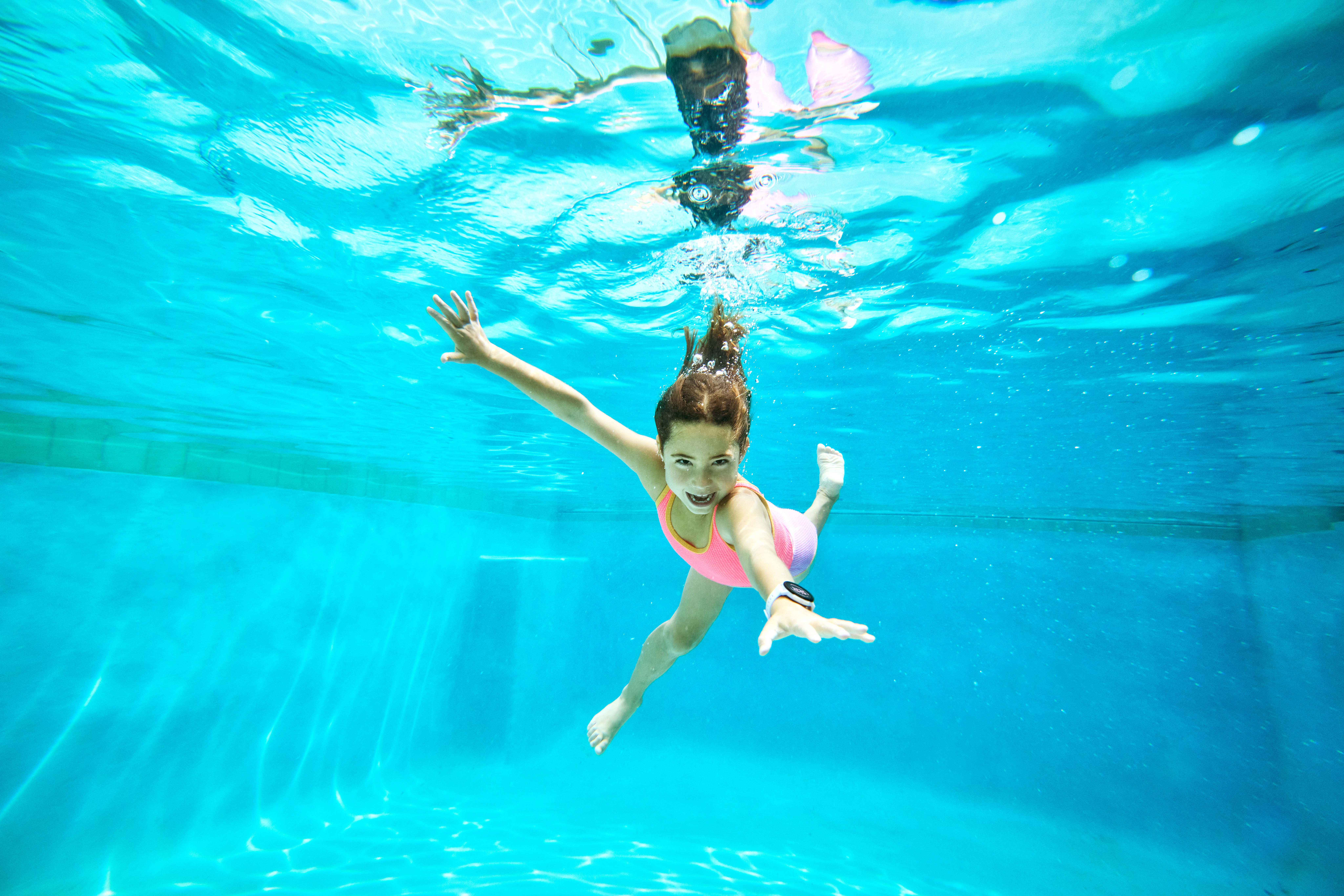 Ein Mädchen schwimmt unter Wasser in einem Pool, die Arme ausgestreckt. Sie trägt einen rosa Badeanzug und lächelt. Licht schimmert durchs Wasser.