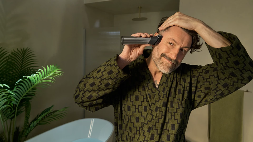 Ein Mann im Bademantel schneidet sich mit einem Haarschneider die Haare. Im Hintergrund sind eine grüne Pflanze und eine weiße Badewanne zu sehen.