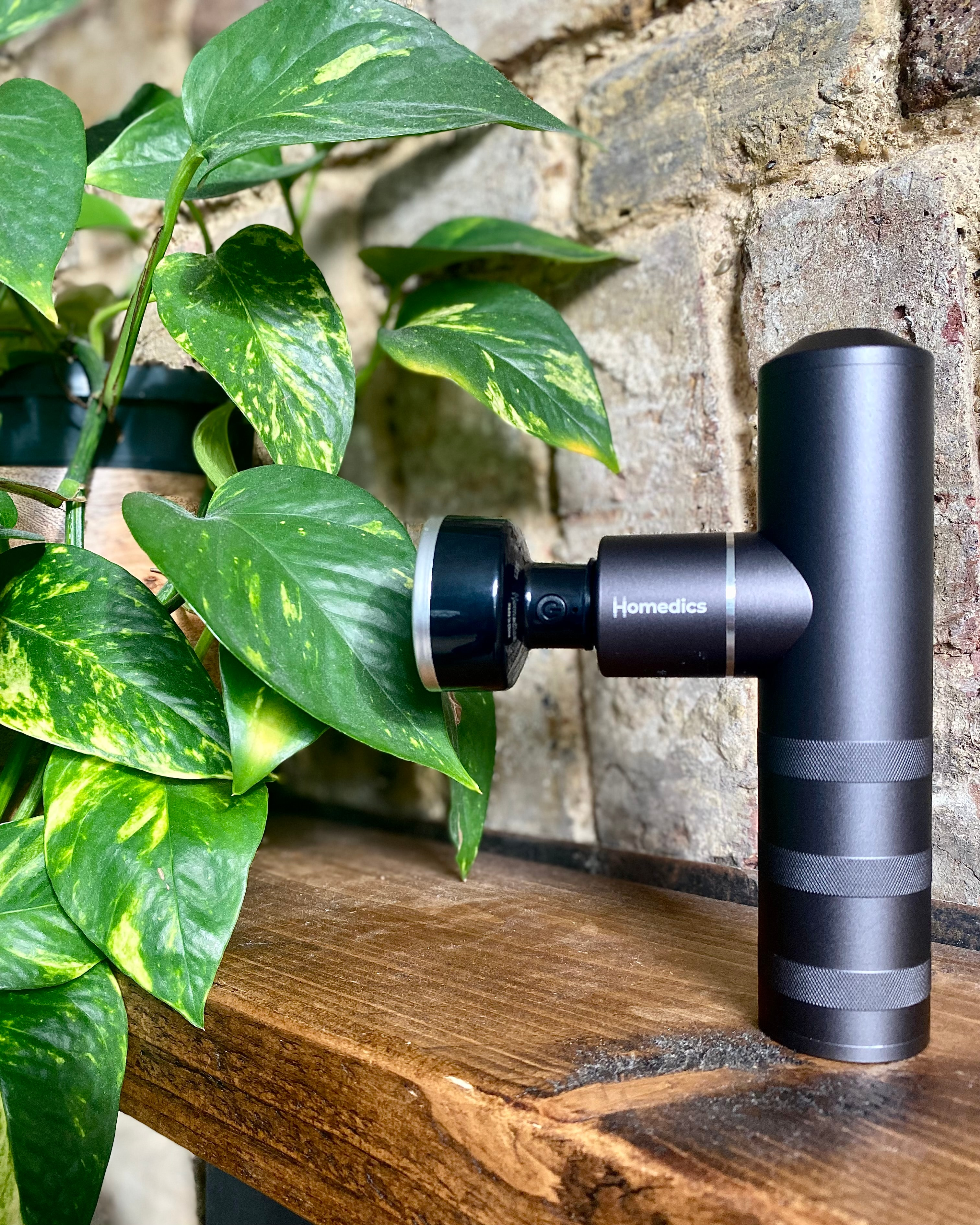 Czarny masażer z zielonymi liśćmi i ceglanym tłem. Urządzenie stoi na drewnianej półce.