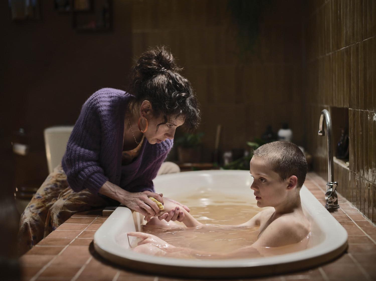 Eine Frau in einem lila Pullover wäscht ein Kind in einer Badewanne. Das Badewasser ist trüb, der Schauplatz ist ein Badezimmer.