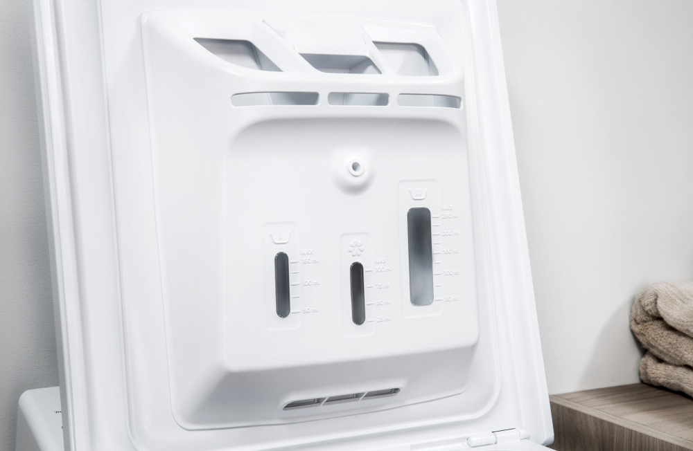 Primo piano del dispenser di detersivo bianco della lavatrice. Ha tre scomparti con segni di misurazione. Asciugamani sullo sfondo.