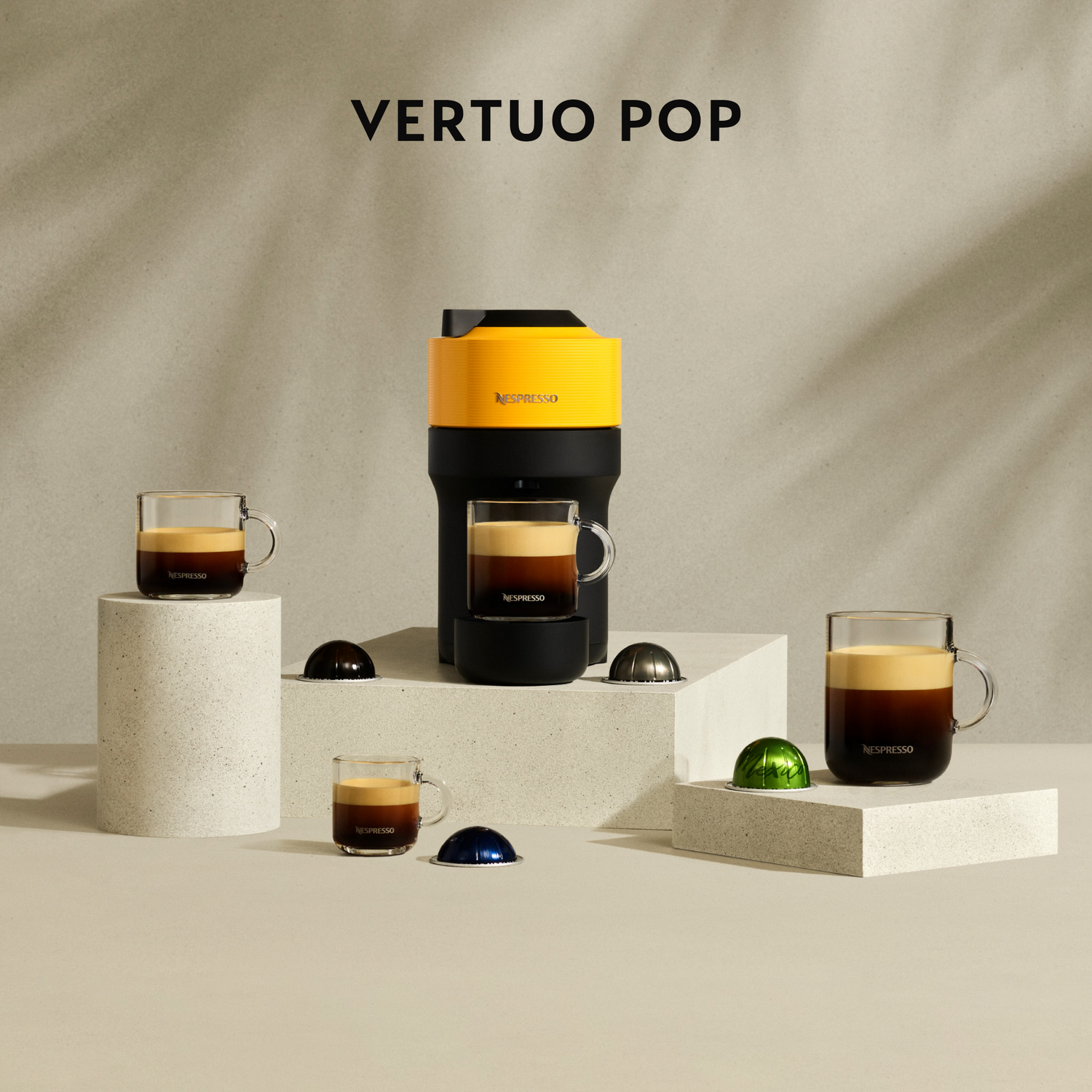 Máquina Nespresso Vertuo Pop con tazas de café, cápsulas y plataformas. Los colores incluyen amarillo, negro, marrón y transparente. Las sombras se proyectan al fondo.