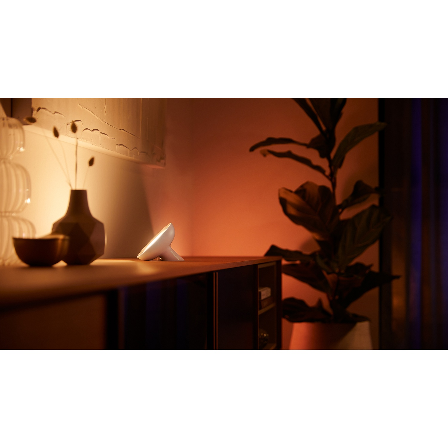 Una lampada bianca su una superficie di legno, con un vaso, una ciotola e una pianta in vaso sullo sfondo. La scena è illuminata da una luce calda.