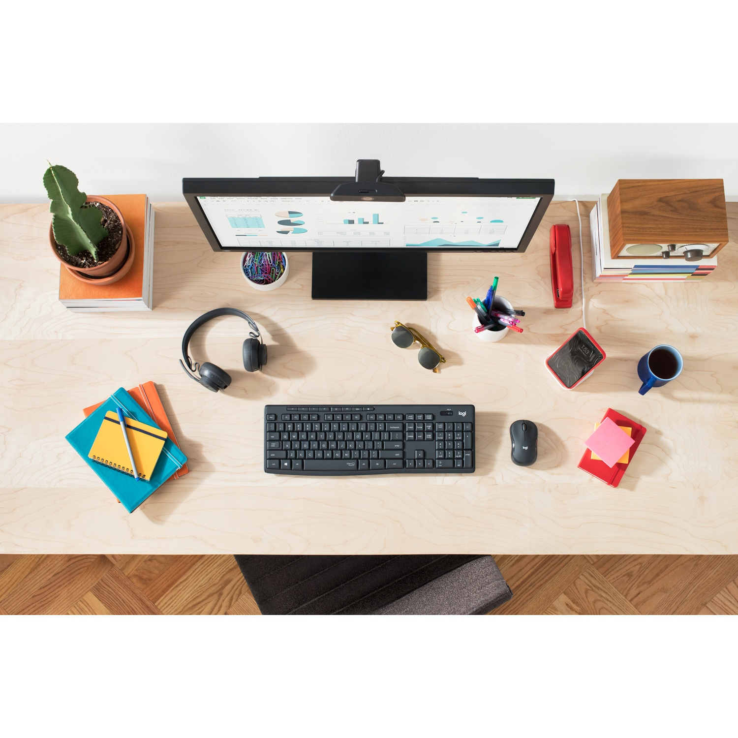 Vista dall'alto di una scrivania: monitor, tastiera, mouse, cuffie, cactus, libri, penne, occhiali da sole, telefono, tazza di caffè e quaderno.