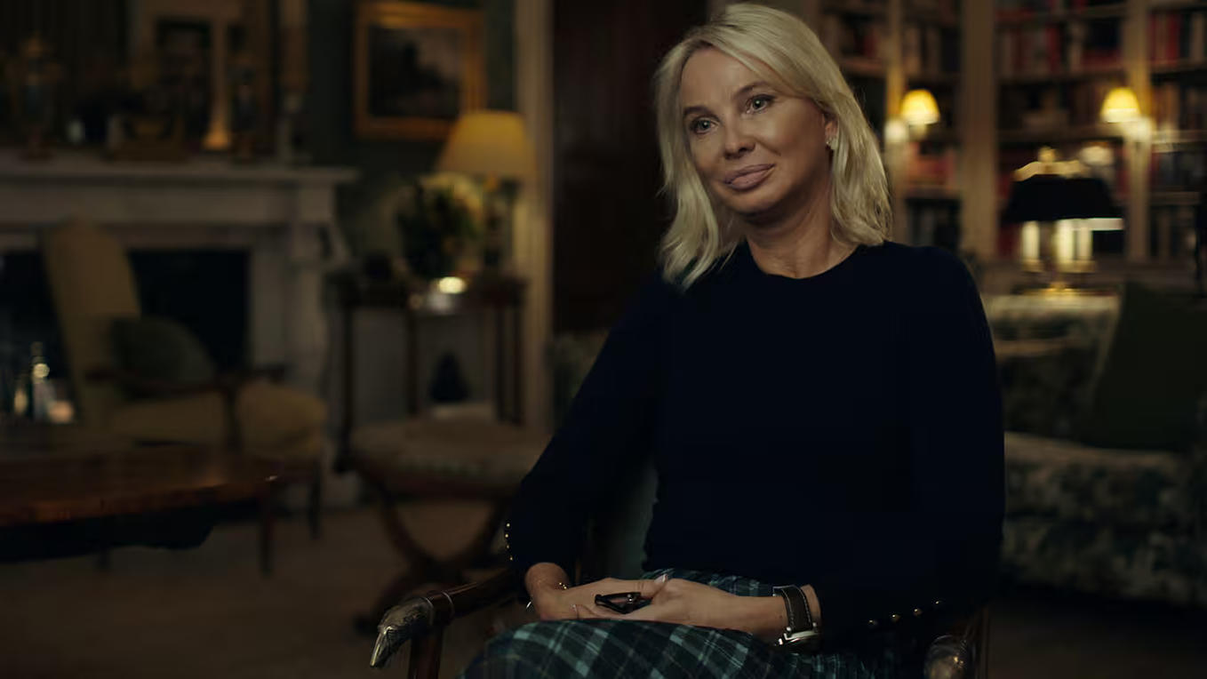 Eine Frau mit blonden Haaren sitzt in einem Raum. Sie trägt einen dunkelblauen Pullover und einen karierten Rock. Im Hintergrund befinden sich ein Kamin und ein Bücherregal.