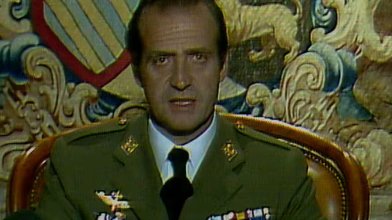 Mann in Militäruniform spricht. Er hat kurze Haare und sitzt. Hinter ihm ein Wandteppich mit Blumenmustern und ein Stuhl.