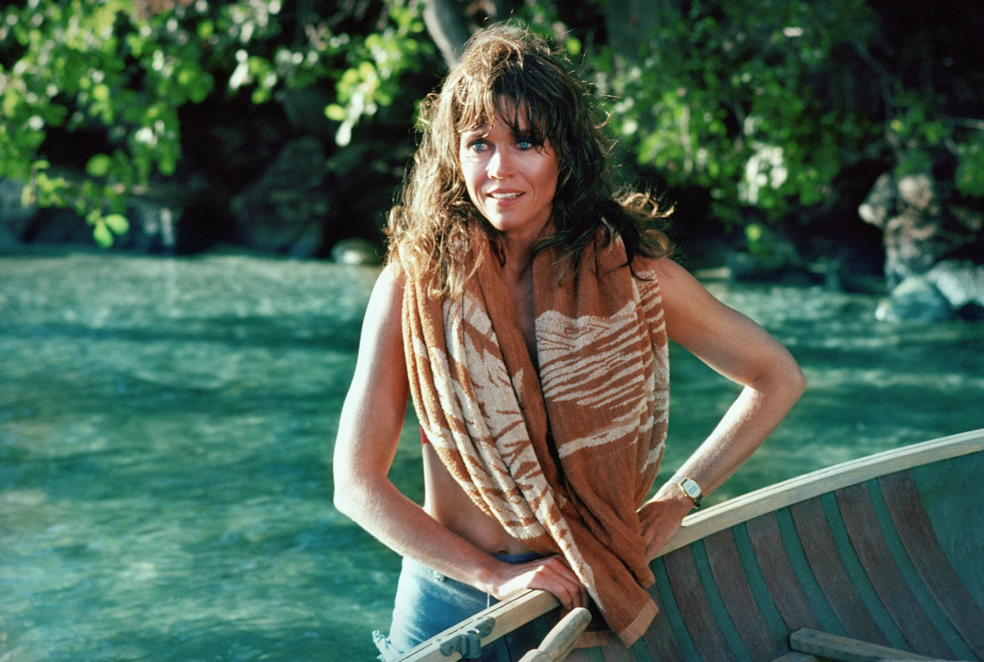 Eine Frau steht in der Nähe eines Bootes, trägt ein Handtuch und blickt in die Kamera. Der Hintergrund ist grünes Wasser und Laub.