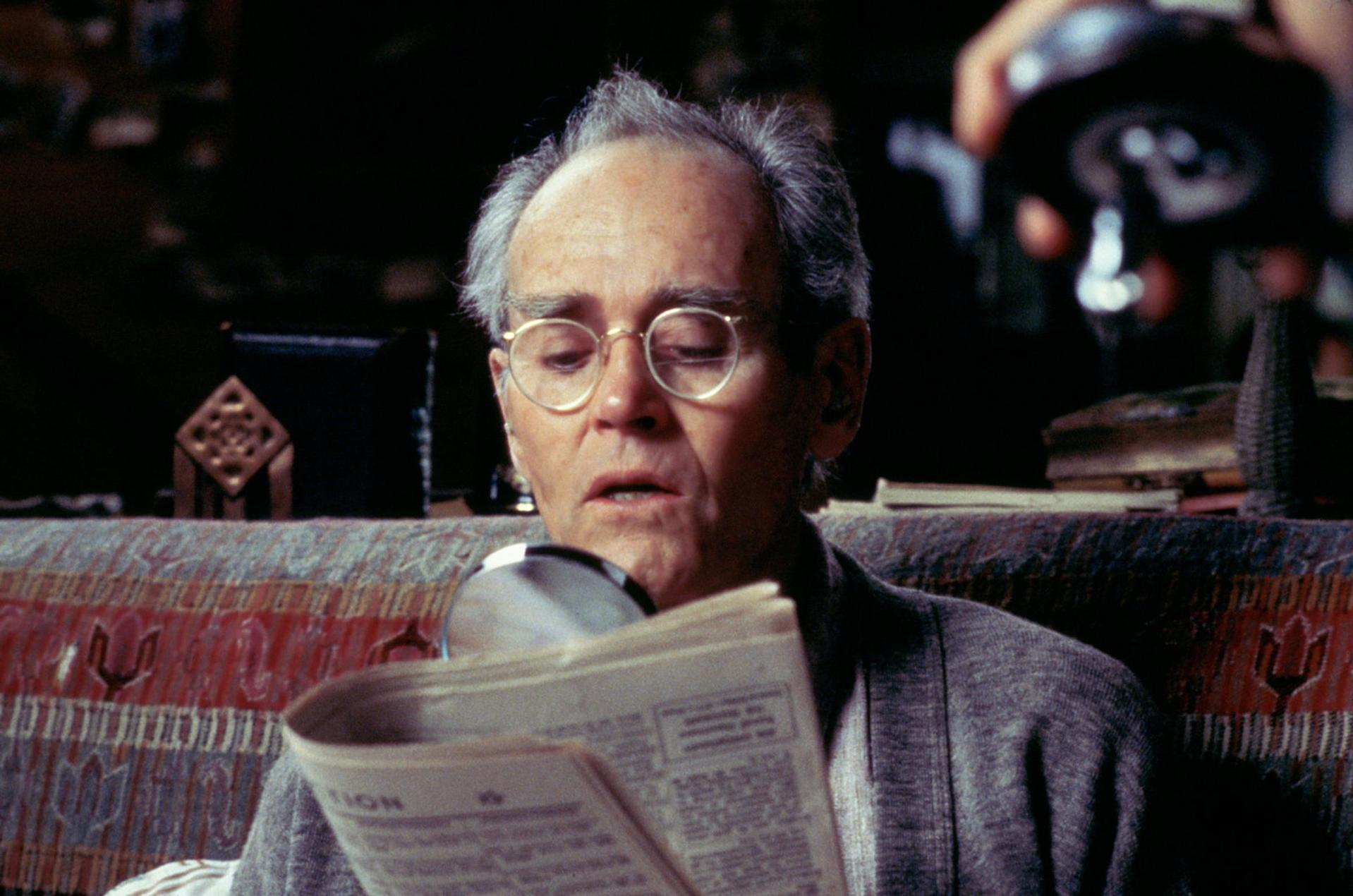 Ein älterer Mann mit Brille liest eine Zeitung, eine Lupe befindet sich in der Nähe des Papiers. Er trägt einen grauen Pullover und sitzt auf einem gemusterten Sofa.