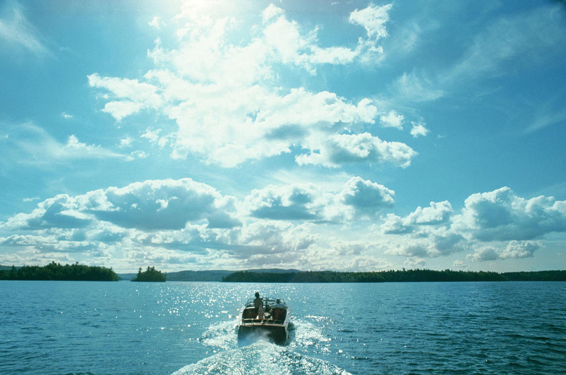 Ein Boot auf einem See mit zwei Personen. Heller Himmel mit Wolken. Bäume am Horizont. Wasser glitzert.