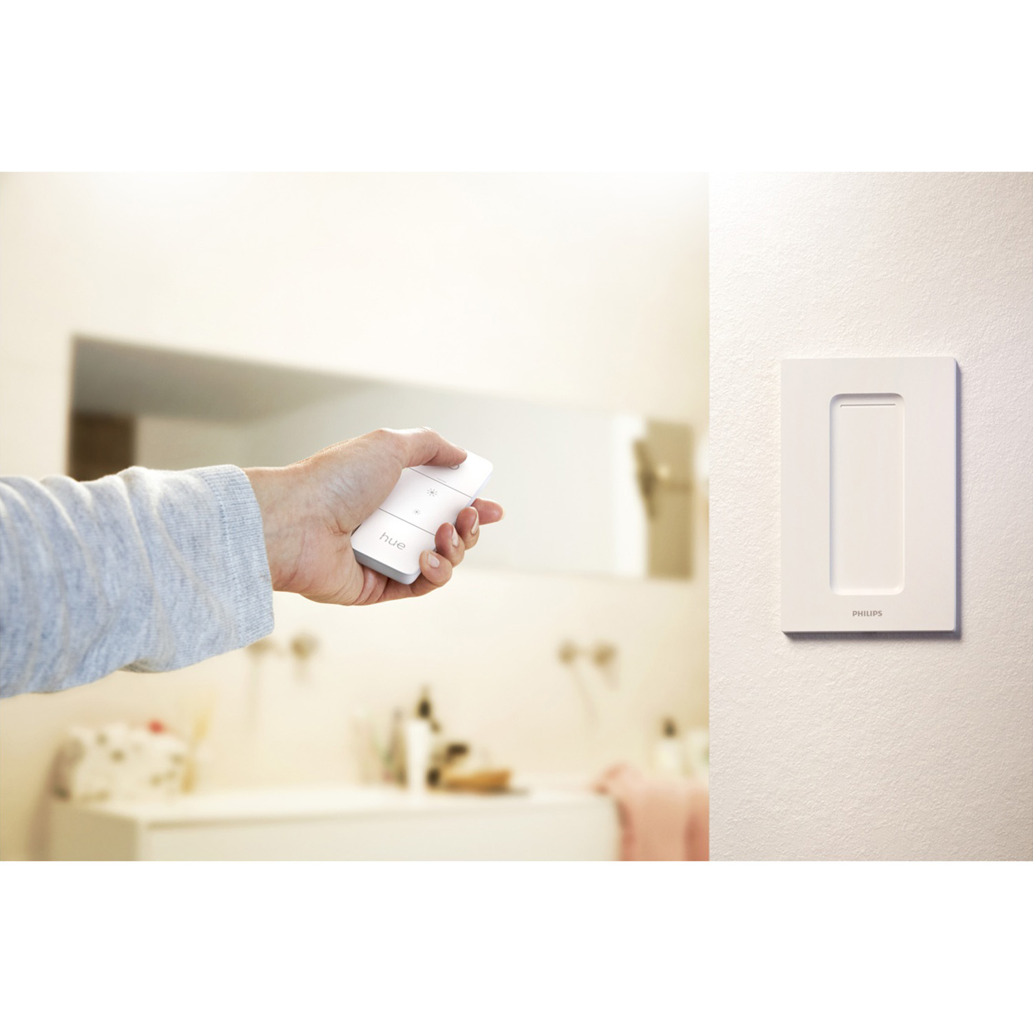 Una mano che tiene un telecomando bianco con la parola 'hue'. Un interruttore della luce è a destra. Una scena di bagno sfocata sullo sfondo.