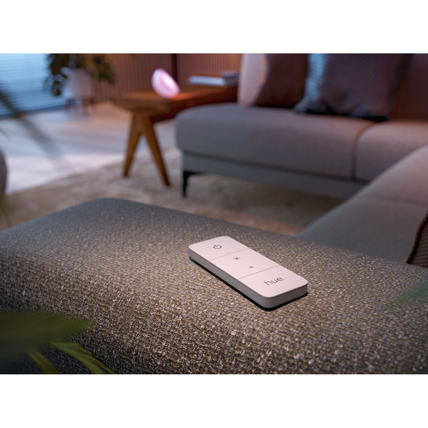 Un telecomando Hue bianco è appoggiato su un pouf grigio strutturato. Un divano e una lampada sono sullo sfondo.