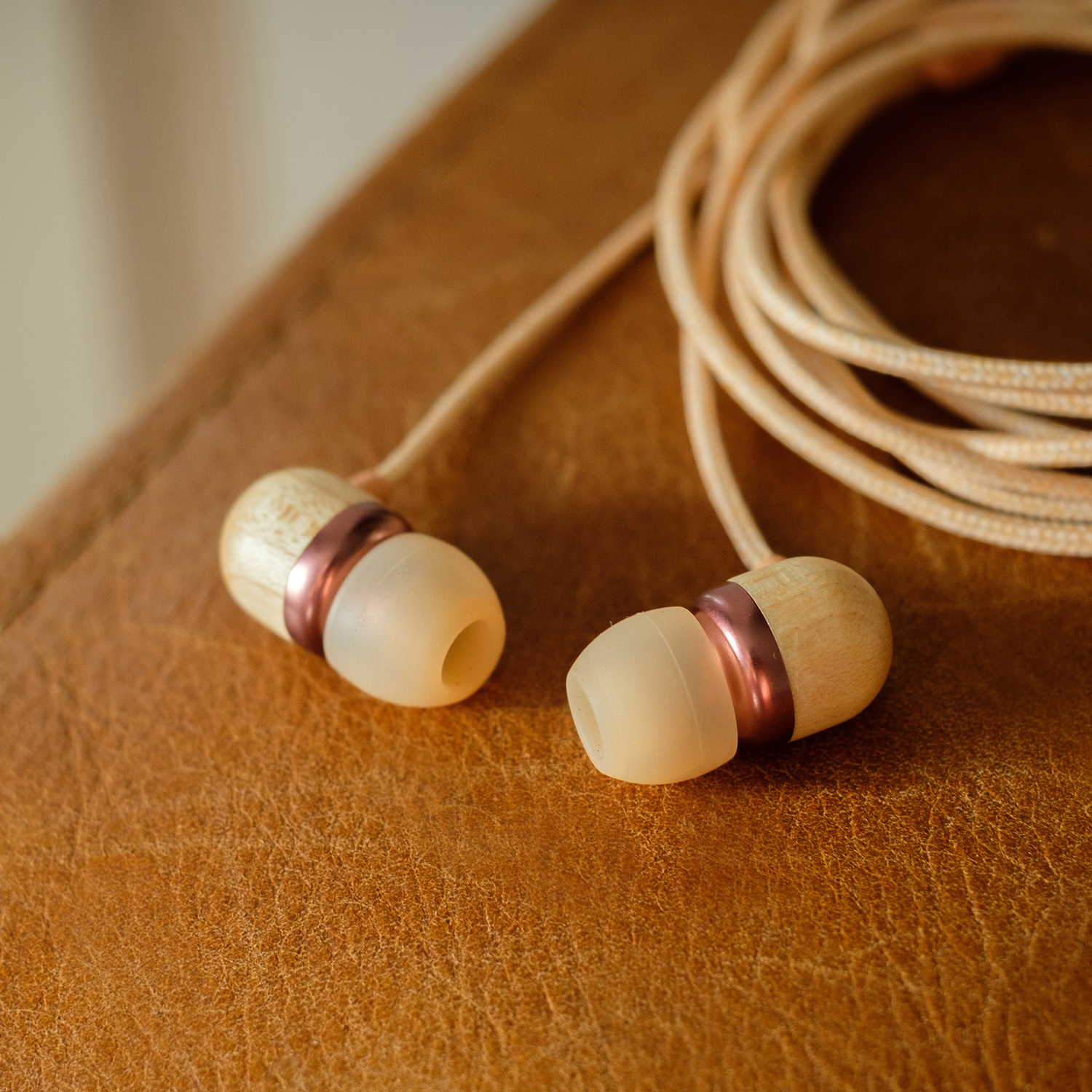 Earbuds with wooden and copper accents. Beige cable coiled on a brown leather surface. Soft focus background.