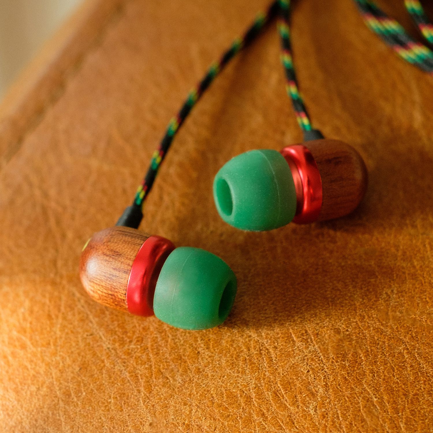 Earbuds on a leather surface. They have wooden casings, red rings, green tips, and multicolored cords. The background is brown leather.