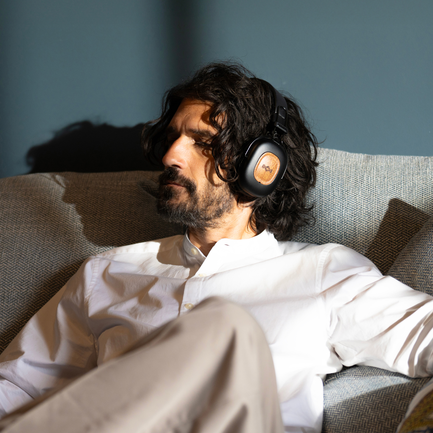 A man with headphones sits on a couch. He wears a white shirt and beige pants. The background is a blue wall.