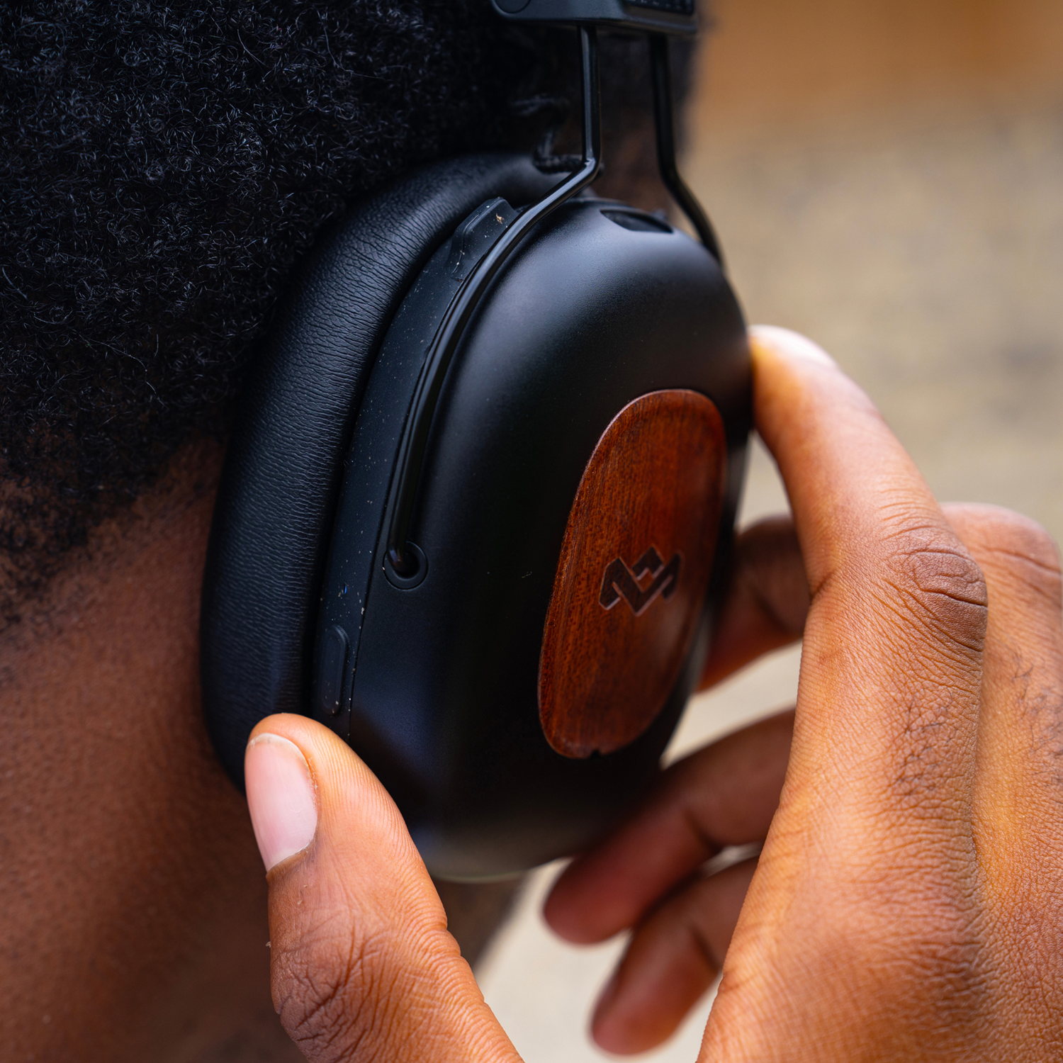 Close-up of a person wearing black headphones with wooden accents. A hand adjusts the headphone. Curly hair is visible.