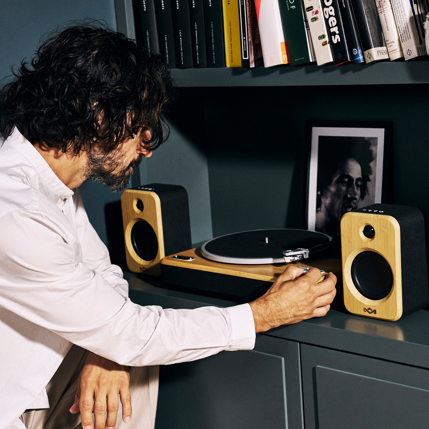 Man die een platenspeler afstelt. Twee luidsprekers met houten fronten op een kast. Boeken op een plank op de achtergrond.