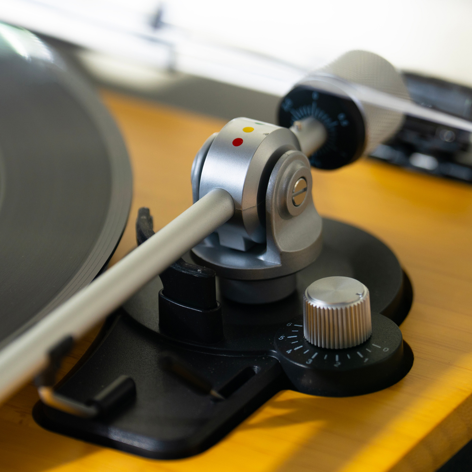 Close-up of a turntable. The silver arm and control knob are visible, with a black record on a yellow wooden surface.