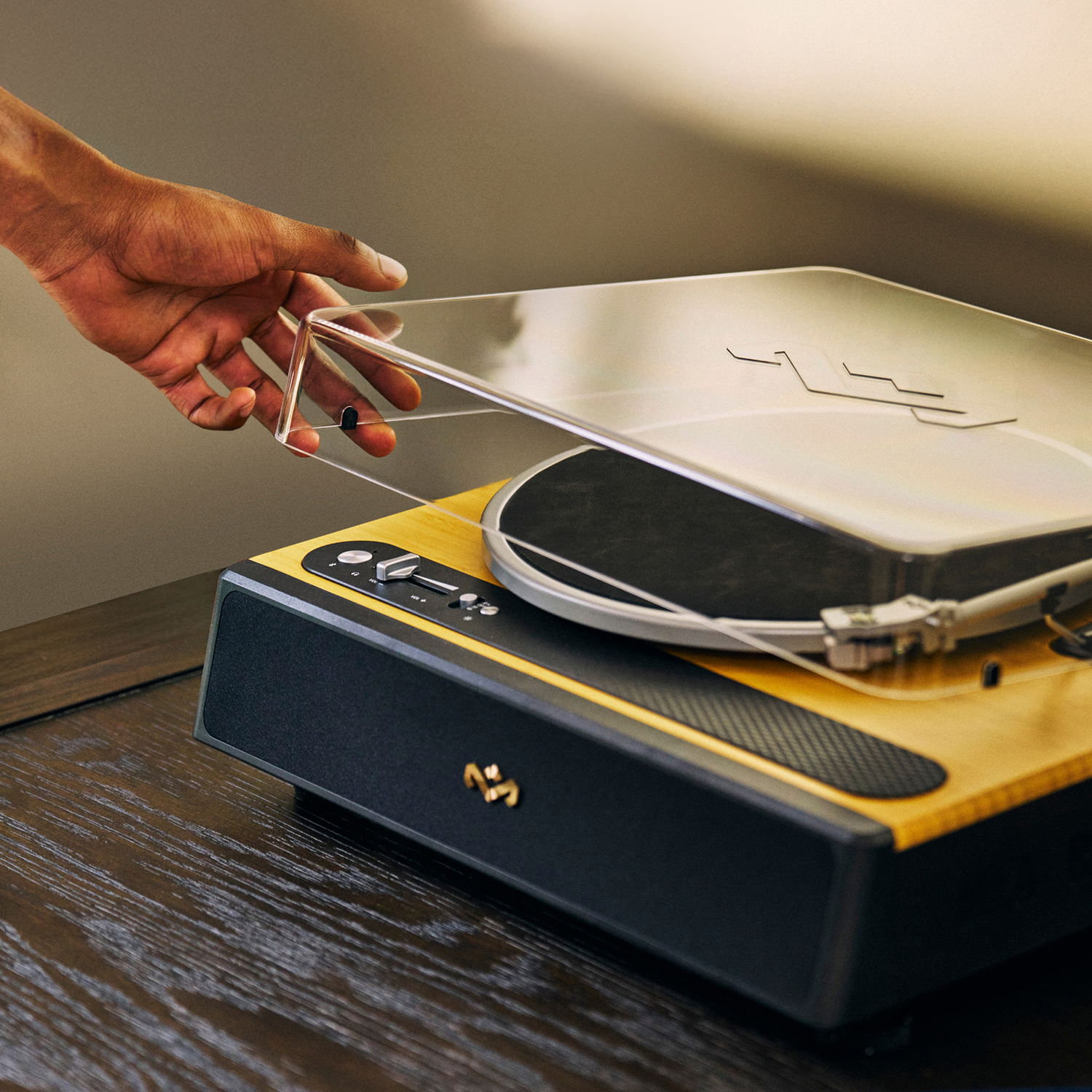 A hand opening the clear cover of a turntable. The turntable is wooden and black, sitting on a dark wooden surface.