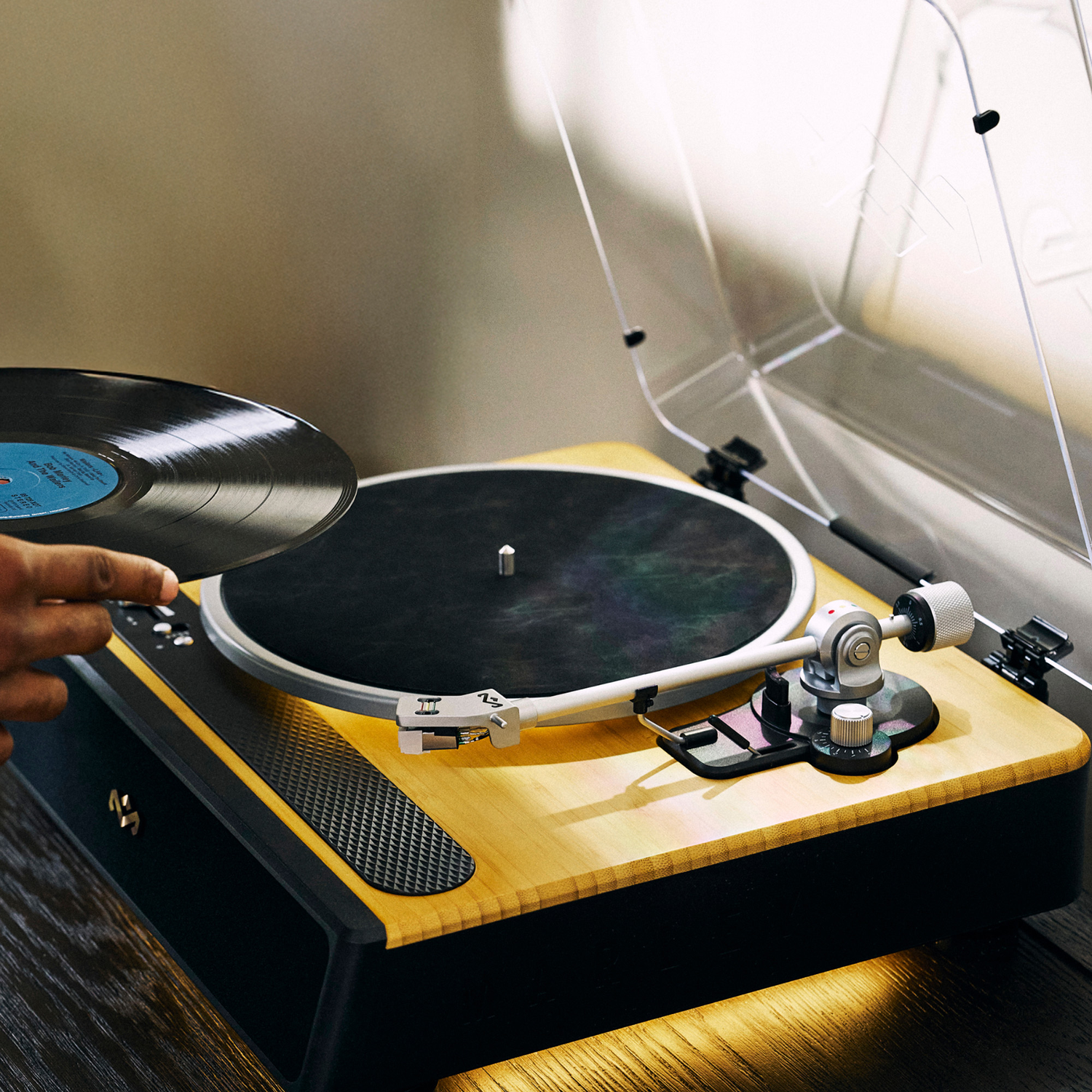 A person places a vinyl record on a wooden record player. The player has a clear cover. The background is a neutral wall.
