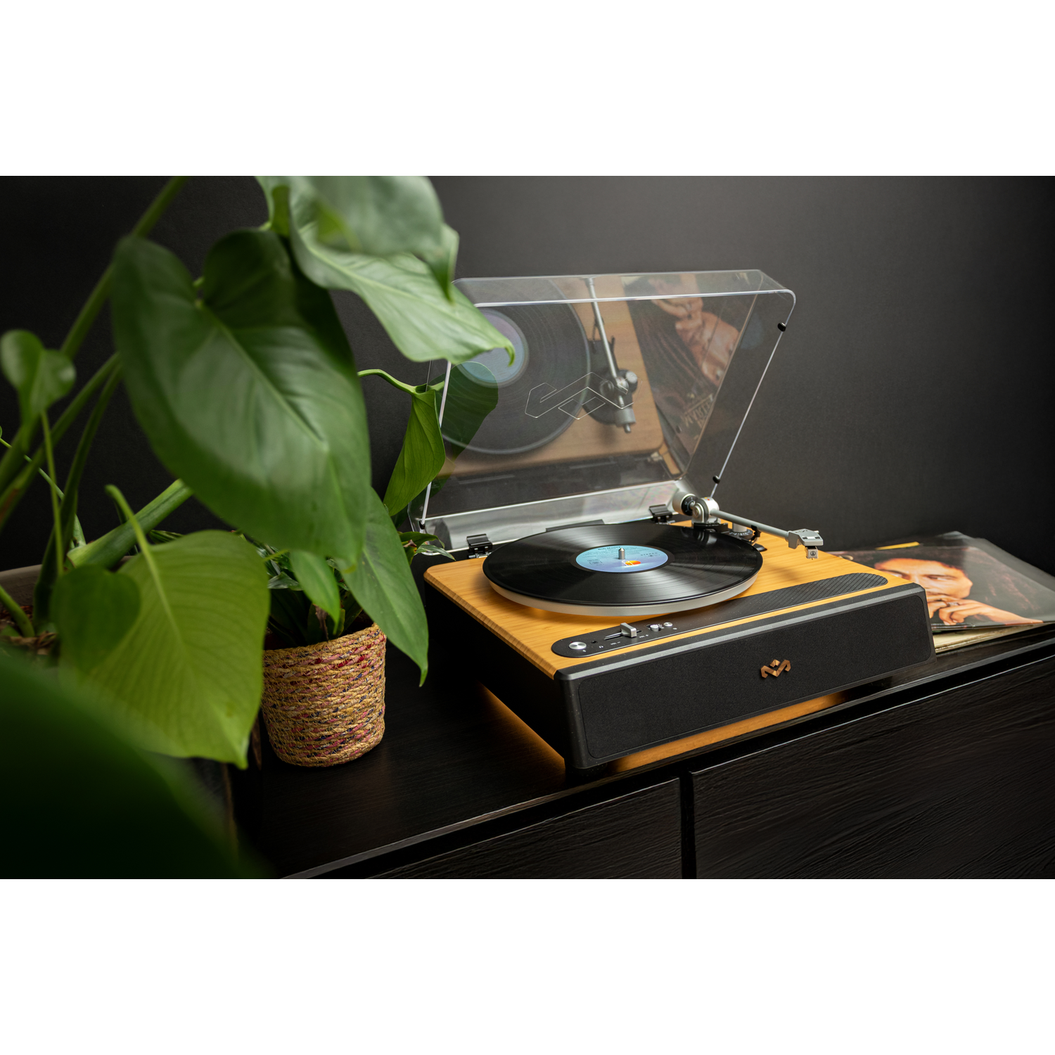 A record player on a dark wooden surface. A vinyl record spins. A potted plant is on the left. A record sleeve is beside the player.