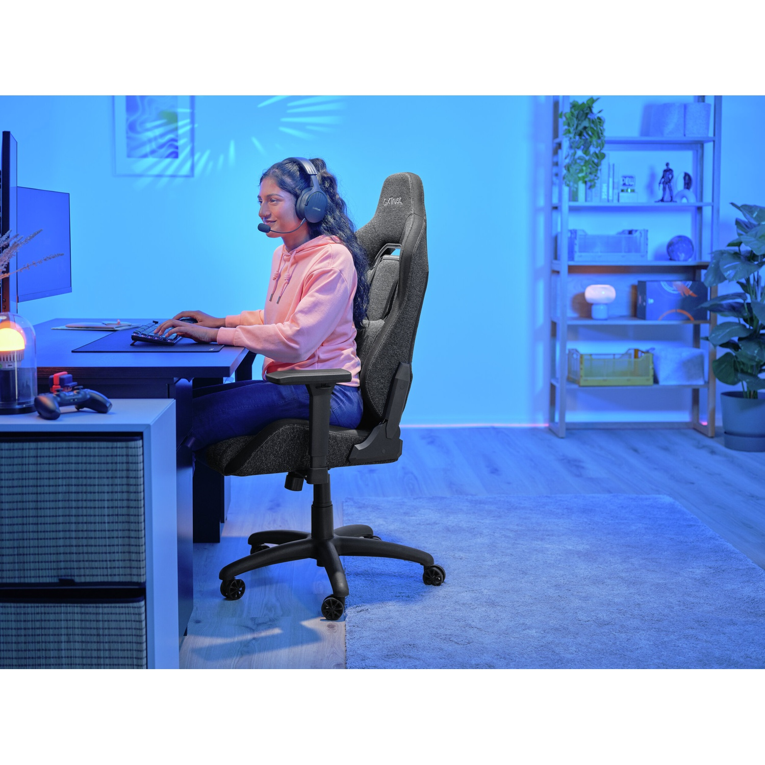 Una donna con le cuffie è seduta a una scrivania, digitando su una tastiera. Una sedia da gioco la sostiene. Un monitor, un controller e delle mensole sono sullo sfondo.