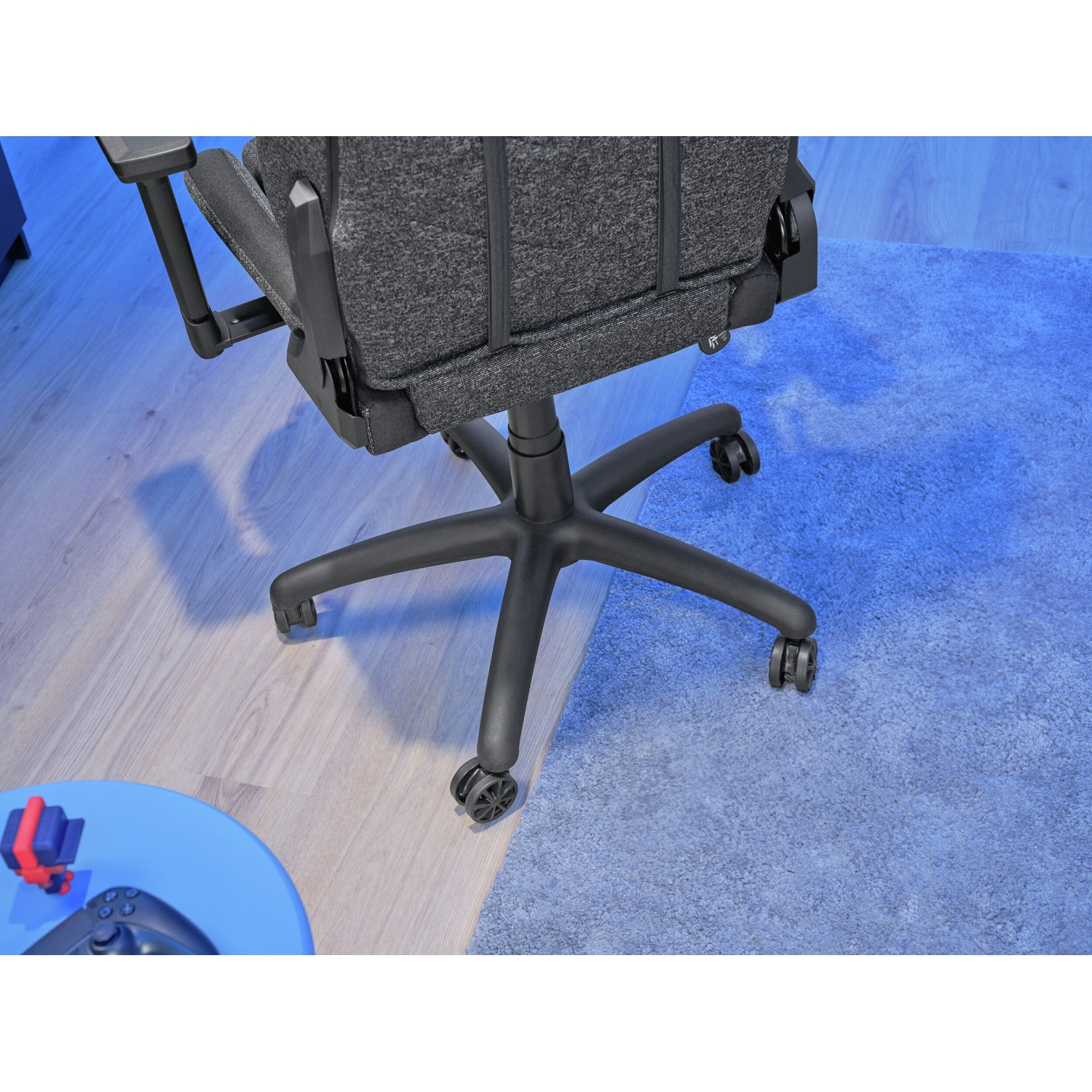 Schienale di una sedia da ufficio grigia con ruote. La sedia è su un pavimento in legno e un tappeto blu. Un tavolo blu con un controller di gioco è visibile.
