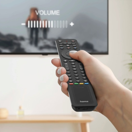 Ręka trzyma czarnego pilota, regulując głośność w telewizorze. Ekran pokazuje rozmyty krajobraz górski.