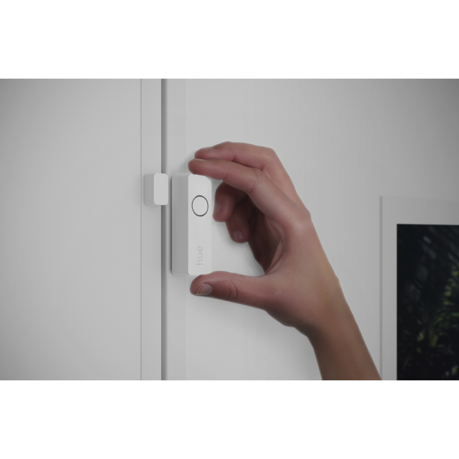 Una mano tiene in mano un dispositivo rettangolare bianco con la scritta 'hue'. Un altro dispositivo è attaccato a un telaio di una porta bianca.
