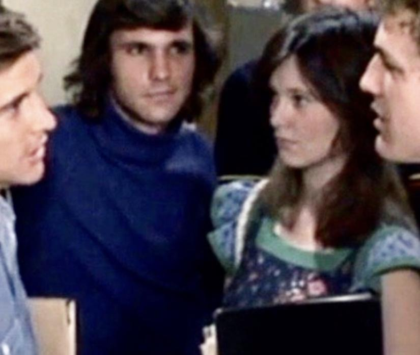 Mehrere Personen befinden sich in einem Raum. Ein Mann in einem blauen Rollkragenpullover steht in der Mitte, neben ihm eine Frau im Blumenkleid.