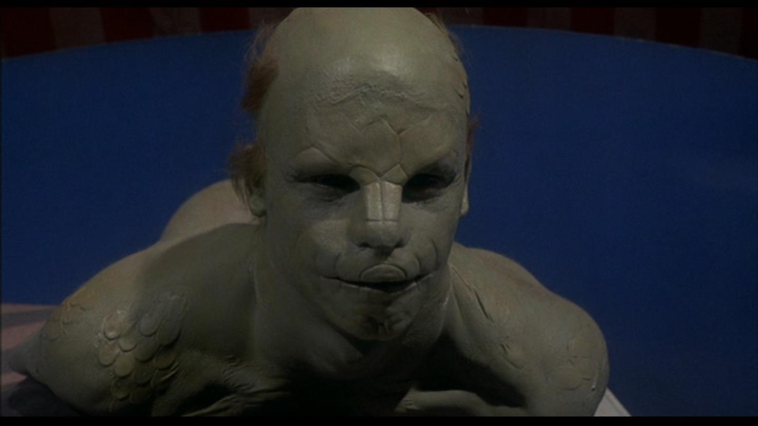 Eine humanoide Figur mit grüner Haut und einem reptilienartigen Gesicht. Der Hintergrund ist blau, mit einer gestreiften rot-weißen zeltartigen Struktur.