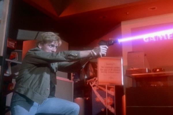 Ein Mann zielt mit einer futuristischen Waffe und feuert einen violetten Laserstrahl auf ein 'SPIELE'-Schild ab. Die Szene spielt in einer schwach beleuchteten Spielhalle.