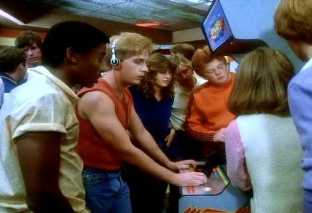 Mehrere Leute um ein Arcade-Spiel. Ein Mann mit Kopfhörern spielt. Andere schauen zu. Der Bildschirm zeigt eine farbenfrohe Spielgrafik.