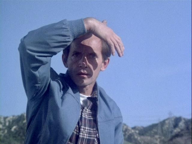 Ein Mann in einer blauen Jacke schützt seine Augen vor der Sonne. Im Hintergrund ist eine Bergkette vor blauem Himmel zu sehen.