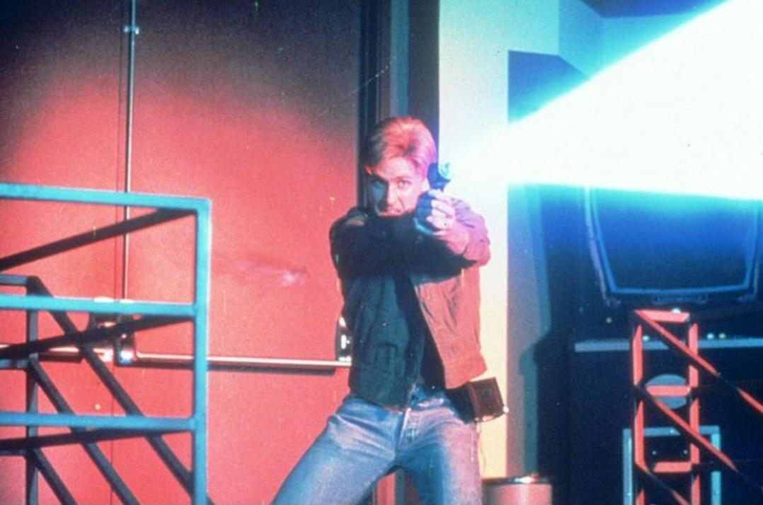 Ein Mann zielt mit einer futuristischen Waffe mit blauem Strahl in einem rot beleuchteten Raum. Er trägt Jeans und eine Jacke und schaut in die Kamera.
