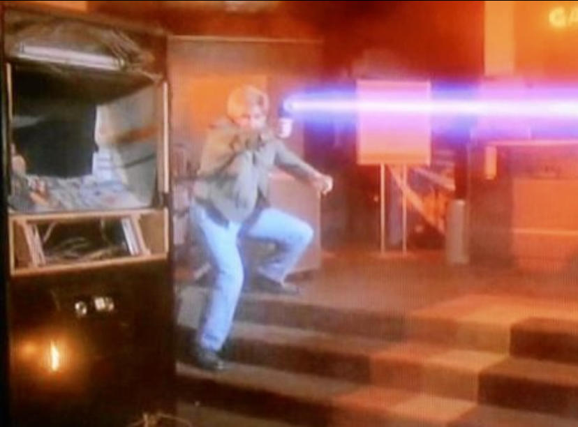 Ein Mann in Jeans und Jacke geht die Treppe hinunter. Ein lila Laserstrahl schießt über die Szene. Spielautomat im Hintergrund.