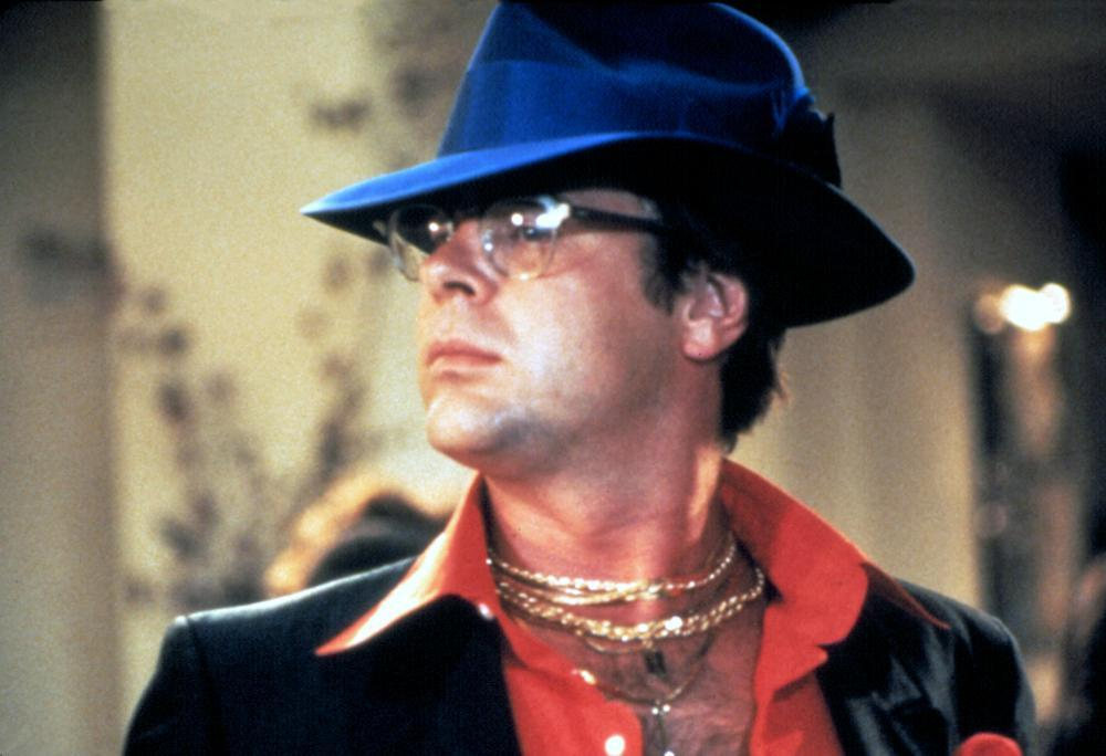 Mann mit blauem Hut, Brille und rotem Hemd mit Goldketten. Er trägt eine schwarze Jacke. Der Hintergrund ist verschwommen.