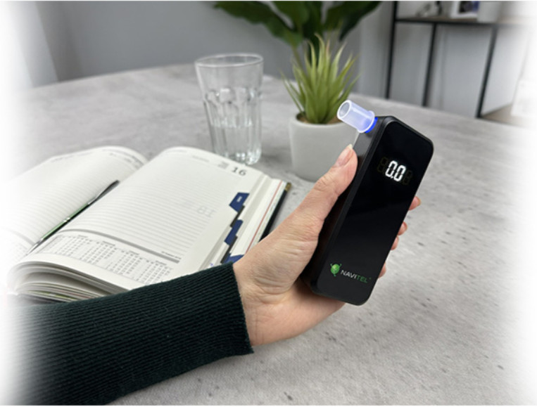 Osoba trzyma czarny alkomat. Szklanka wody, roślina i otwarty notatnik leżą na stole. Odczyt wynosi 0,0.