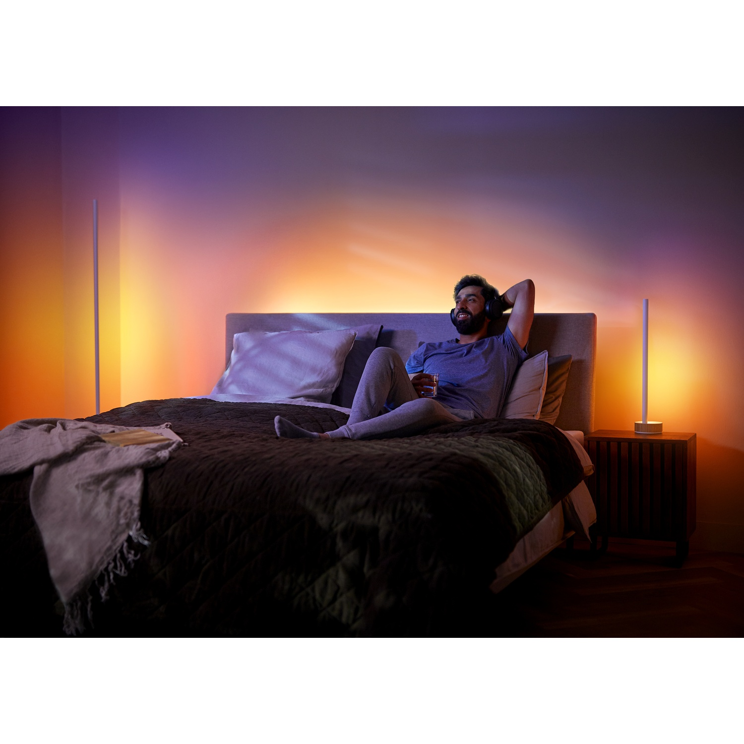 Un uomo con le cuffie si rilassa su un letto. Tiene un bicchiere, due lampade su entrambi i lati. Lo sfondo ha sfumature arancioni e viola.