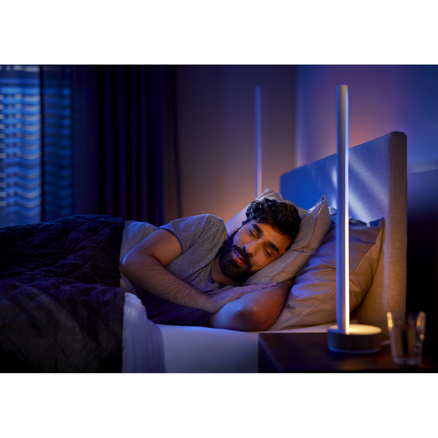 Uomo addormentato a letto con una lampada da comodino. Stanza buia con sfumature blu e viola. L'uomo indossa una maglietta grigia.