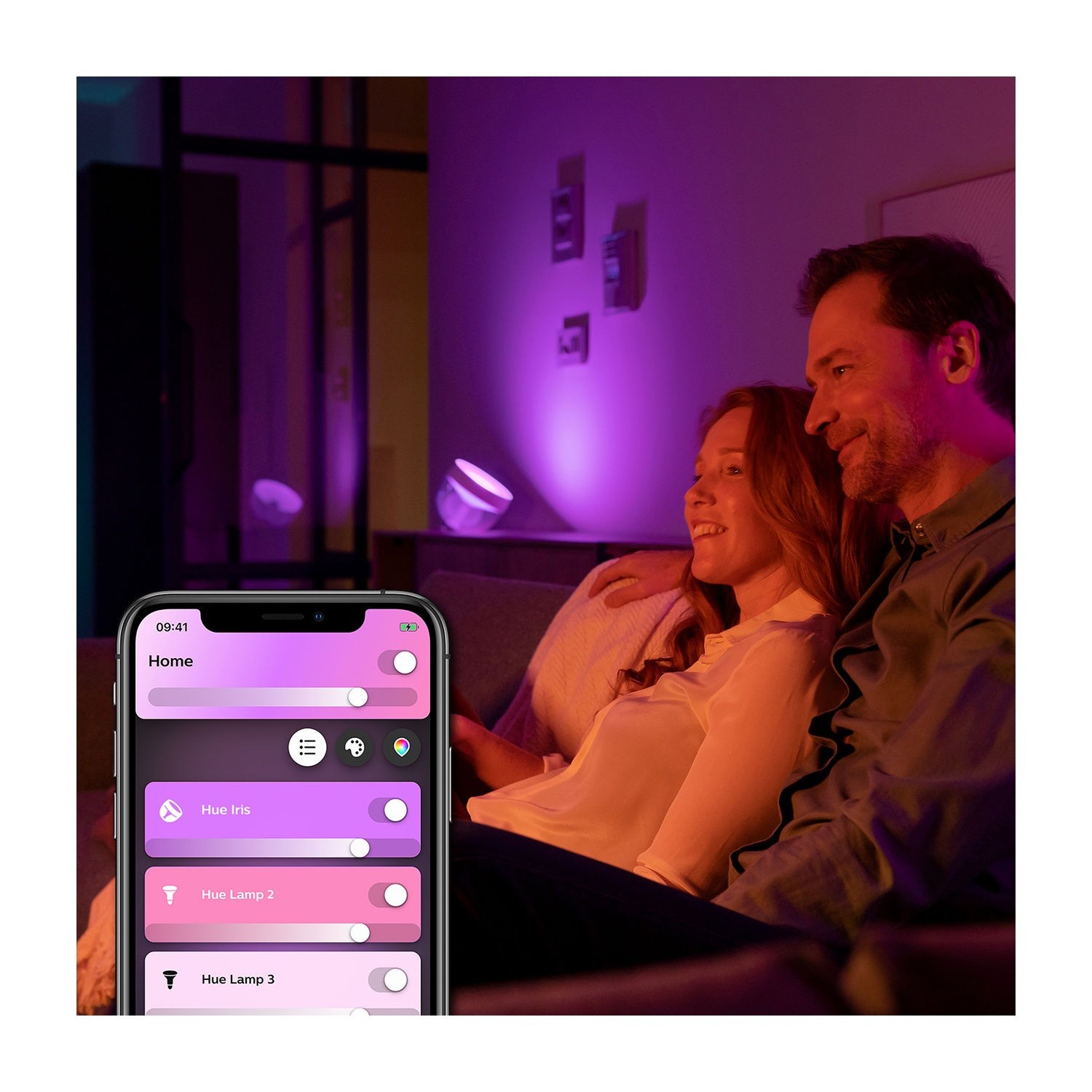 Una coppia seduta su un divano in una stanza illuminata da luci viola e arancioni, che utilizza un telefono per controllare le luci.