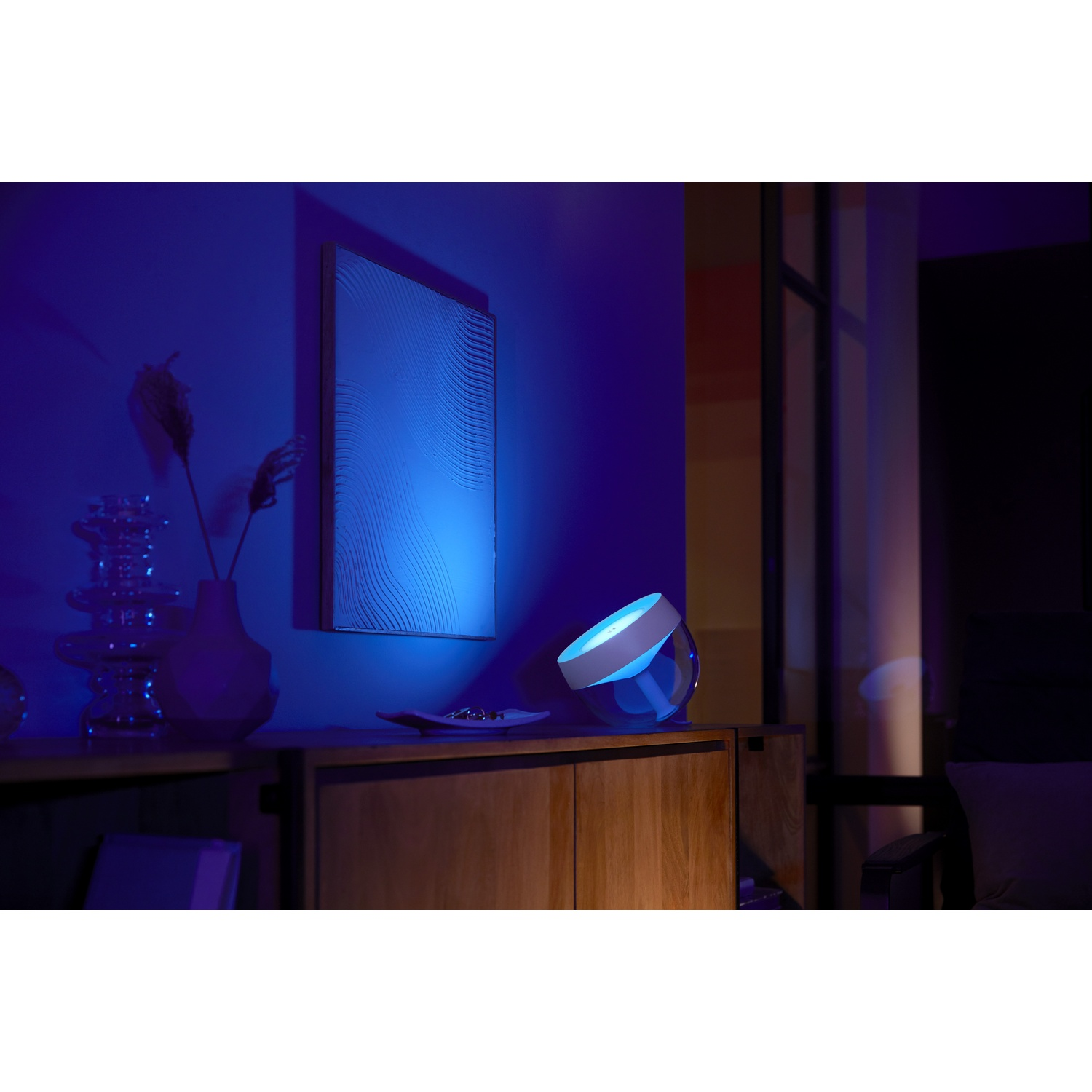 Una lampada blu su un mobile in legno. Un quadro appeso al muro. Un vaso con piante è anche sul mobile.