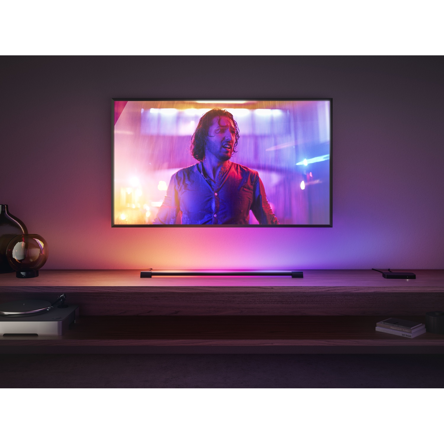 Un uomo sullo schermo di una TV con luci colorate. Una mensola in legno ospita un giradischi e una barra luminosa. Il muro è scuro.