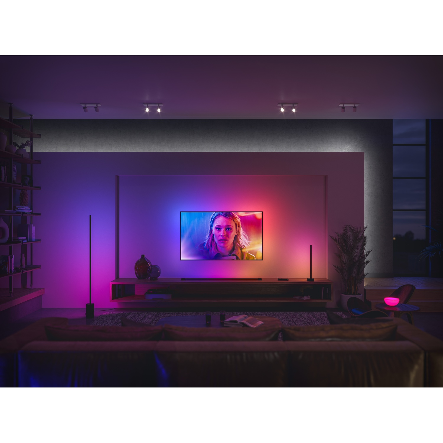 Un soggiorno con una grande TV che mostra il volto di una donna. La stanza è illuminata da luci colorate e un divano marrone. Due luci nere.