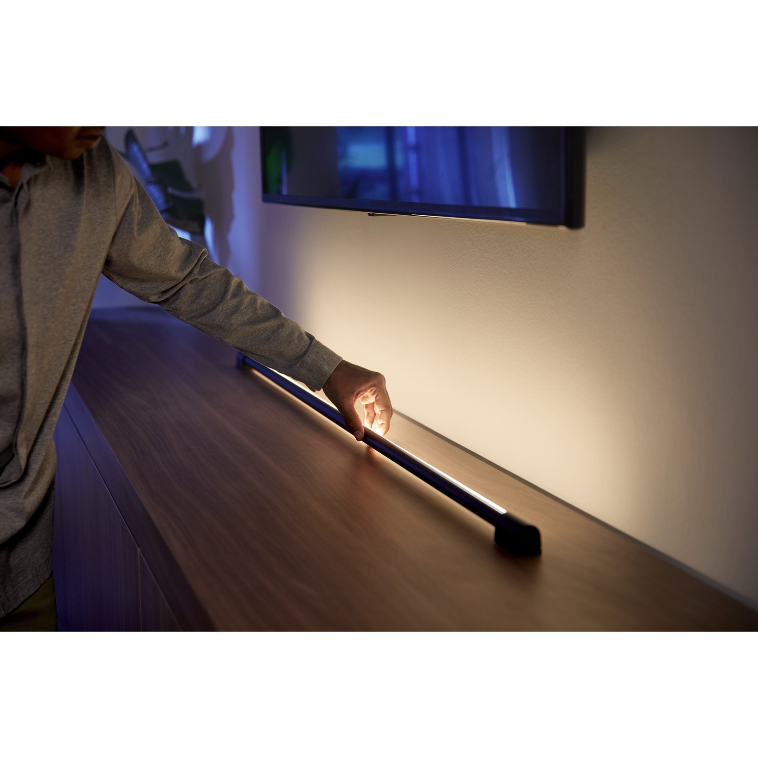 La mano di una persona tocca una barra luminosa nera su una superficie di legno. C'è una TV sullo sfondo e il muro è illuminato.