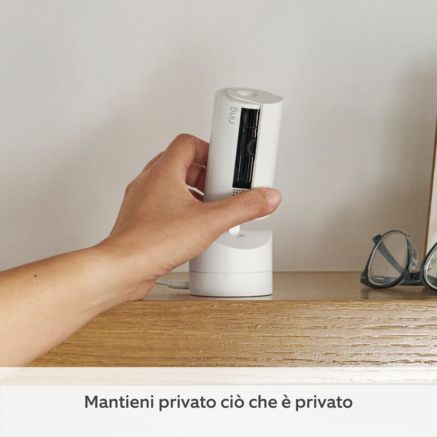Mano che tiene una telecamera di sicurezza bianca su una mensola di legno. Occhiali sulla mensola. Testo in basso: Mantieni privato ciò che è privato.