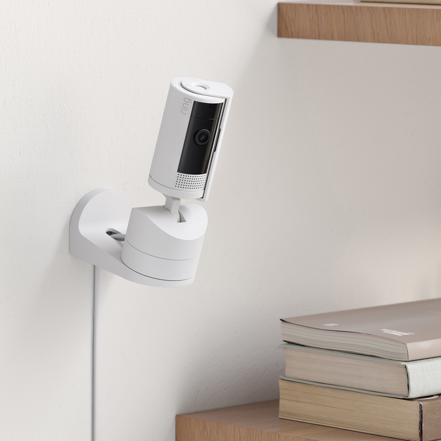 Una telecamera di sicurezza Ring bianca montata a parete. Libri su una mensola di legno. Un cavo corre lungo il muro.